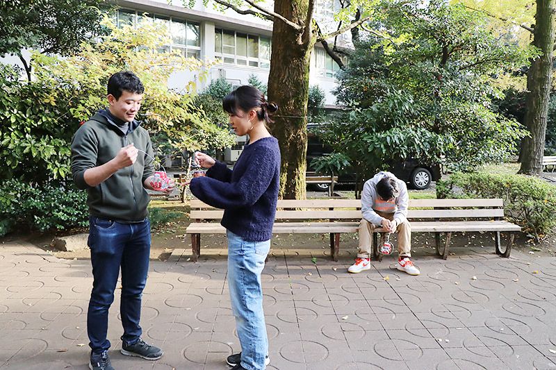 空き缶風車を見せ合う大学生2名と、疲れて再びベンチに座る地主